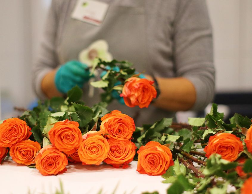 A professional florist from Floristroehampton.co.uk arranges a floral bouquet featuring vibrant orange roses with lush green foliage, displayed on a white work surface. The florist, wearing a grey top