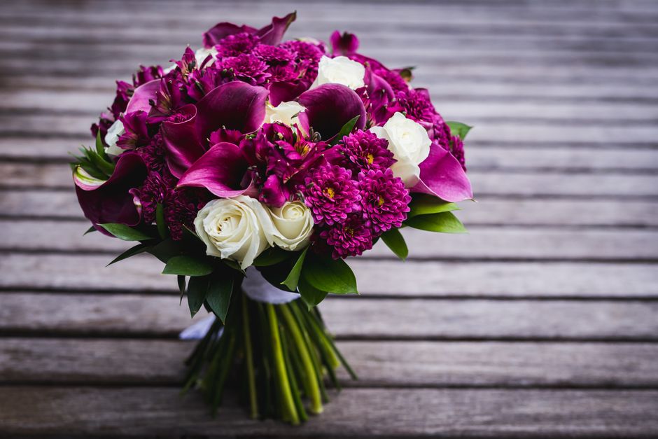 A bouquet of fresh flowers placed on a wooden surface, featuring deep purple calla lilies, magenta chrysanthemums, purple roses, and small white roses. The arrangement is wrapped with white ribbon, wi