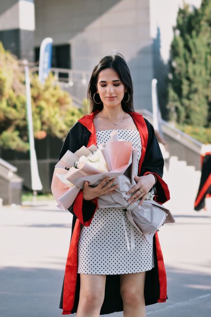A young woman with long dark hair, dressed in a white polka dot dress and a black graduation gown with red lining, is holding a bouquet of fresh flowers wrapped in pale pink and white paper with some
